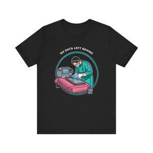 May include: A black t-shirt featuring a graphic of a medical professional in teal scrubs and a face mask working at a computer station. The text NO DATA LEFT BEHIND is printed in white in an arc above the circular illustration of the scene.
