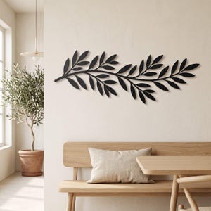 Op de afbeelding: Een matzwart metalen wanddecoratie in de vorm van een lange, horizontale olijftak met talloze puntige bladeren. Het ontwerp heeft een centraal, licht gebogen steeltje met kleinere takjes die naar buiten uitsteken, elk voorzien van meerdere bladeren.