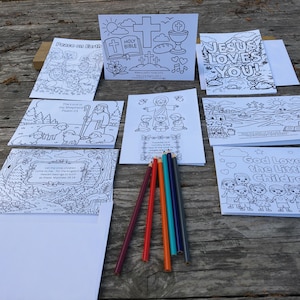 May include: A collection of coloring pages with religious themes, including images of Jesus, crosses, and Bible verses. Several colored pencils in red, orange, and blue hues are arranged on a wooden surface. The pages are blank and ready to be colored.