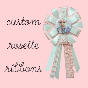 May include: A custom rosette ribbon featuring a circular pin with a portrait of a person, surrounded by layered pale pink and white ribbons. The design includes vertical ribbon tails decorated with small heart patterns and pink trim.