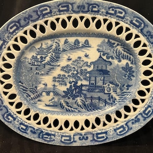 May include: An oval ceramic platter featuring a blue and white willow pattern illustration of a landscape with pagodas, trees, and a bridge. The rim has a decorative openwork lattice design with a repeating geometric border pattern.