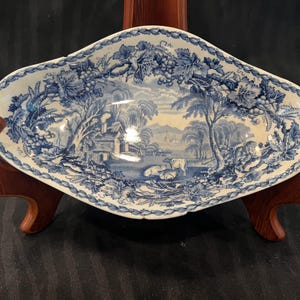May include: An oval ceramic dish featuring a blue and white transferware pattern. The design depicts a pastoral landscape with a cottage, trees, and grazing cattle, framed by a decorative border of grapevines and leaves along the scalloped rim.
