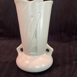 May include: A pale, off-white ceramic vase with a smooth, matte finish. The vase features a flared, tulip-like rim, two small side handles, and a raised, stylized leaf or feather relief pattern on the front with subtle light green accents.