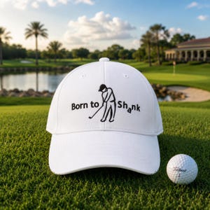 Pode incluir: Um boné de beisebol branco com o texto "Born to Shank" e um gráfico com tema de golfe. Uma bola de golfe está ao lado do boné em um campo de golfe verde. O boné tem um design clássico de seis painéis.