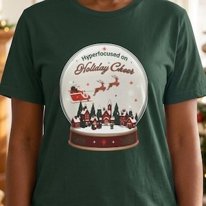 Op de afbeelding: Een donkergroen T-shirt met korte mouwen en een afbeelding van een sneeuwbol. Binnenin de bol staat een winters dorpsgezicht met een rode arrenslee getrokken door rendieren en de tekst "Hyperfocused on Holiday Cheer" in een cursief lettertype.