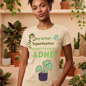 May include: A beige short-sleeved t-shirt featuring a green graphic of a hanging plant, a small watering can, and a potted plant with a smiling face. The text reads: my latest hyperfixation brought to you by ADHD in green lettering.