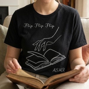 May include: Black t-shirt with the words "Flip Flip Flip" and a line drawing of a hand turning a book page. The word "ASMR" is printed below the book illustration. The person is holding an open book.
