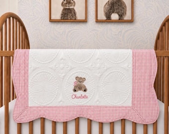 Könnte beinhalten: Eine weiße, gesteppte Babydecke mit einem rosa karierten, gewellten Rand. In der Mitte befindet sich ein gestickter brauner Teddybär mit einer rosa Schleife und dem Namen Charlotte in rosa Garn unter dem Bären.