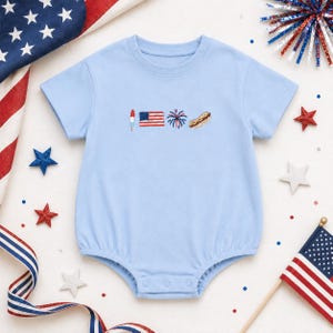 Könnte beinhalten: Ein hellblauer, kurzärmeliger Baby-Body mit einer kleinen Stickerei auf der Brust. Das Motiv zeigt ein rot-weiß-blaues Eis am Stiel, eine amerikanische Flagge, ein blau-rotes Feuerwerk und einen Hotdog in einem Brötchen.