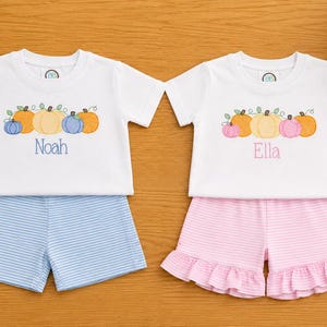 May include: Two white short-sleeve t-shirts paired with shorts. The left set features blue striped shorts and a pumpkin graphic with the name Noah in blue. The right set features pink striped ruffled shorts and a pumpkin graphic with the name Ella in pink.