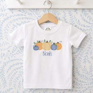 K&ouml;nnte beinhalten: Ein wei&szlig;es kurz&auml;rmeliges T-Shirt mit einer Stickerei von vier K&uuml;rbissen in Orange- und Blaut&ouml;nen mit gr&uuml;nen Ranken. Der Name Noah ist in blauem Garn unter den K&uuml;rbissen eingestickt. Das T-Shirt h&auml;ngt an einem h&ouml;lzernen Kleiderb&uuml;gel.
