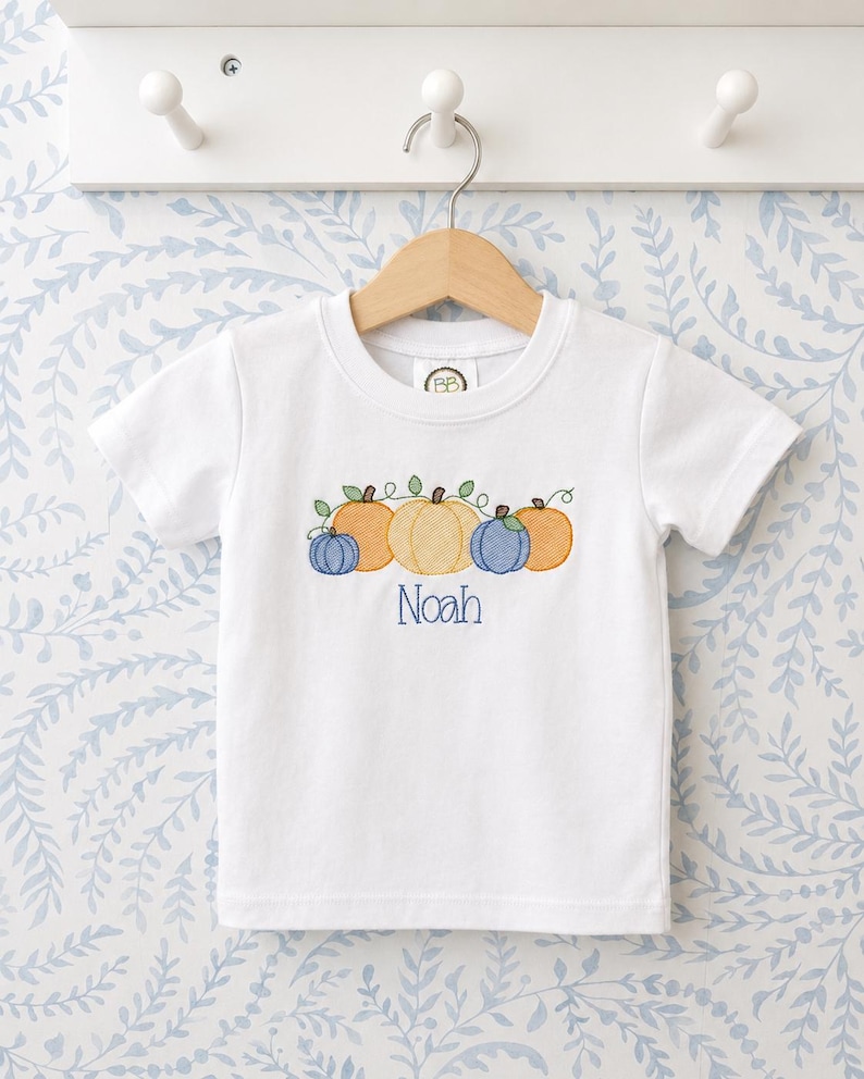 K&ouml;nnte beinhalten: Ein wei&szlig;es kurz&auml;rmeliges T-Shirt mit einer Stickerei von vier K&uuml;rbissen in Orange- und Blaut&ouml;nen mit gr&uuml;nen Ranken. Der Name Noah ist in blauem Garn unter den K&uuml;rbissen eingestickt. Das T-Shirt h&auml;ngt an einem h&ouml;lzernen Kleiderb&uuml;gel.