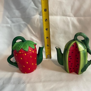 Può includere: Due teiere in ceramica, una a forma di fragola rossa con foglie verdi e l'altra a forma di anguria. La teiera a fragola ha puntini gialli. La teiera ad anguria ha scorza verde e interno rosso.