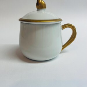 May include: A white ceramic mug with a matching lid, featuring a gold-colored handle, a gold-colored rim on the lid, and a small gold-colored knob on top of the lid. The mug has a rounded, bulbous body shape and a smooth, glossy finish throughout.