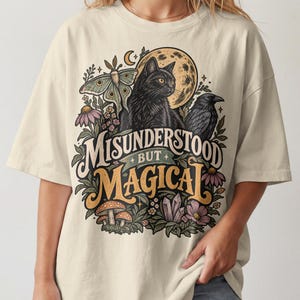 May include: An off-white short-sleeve t-shirt featuring a graphic print of a black cat, a raven, a moth, flowers, mushrooms, and a moon. The text reads MISUNDERSTOOD BUT MAGICAL in a stylized font. The design includes earthy tones of gold, green, and purple.