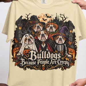 May include: A cream-coloured t-shirt featuring a graphic of five bulldogs dressed in Halloween costumes, including a ghost, a vampire, and a skeleton. The text "Bulldogs Because People Are Creepy" is printed below the dogs, surrounded by pumpkins and skulls.