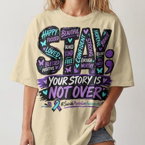 May include: A beige t-shirt featuring a large graphic with the text STAY YOUR STORY IS NOT OVER. The design includes words like HAPPY, FOCUSED, LOVED, BLESSED, POSITIVE, BEAUTIFUL, BALANCED, KIND, FREE, CONFIDENT, STRONG, ENOUGH, WORTHY, HOPEFUL, BRAVE, RESILIENT, YOU MATTER.