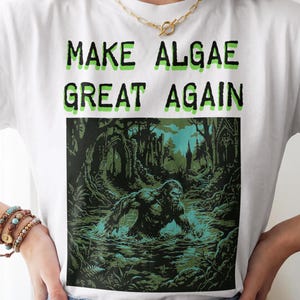 Könnte beinhalten: Ein weißes T-Shirt mit dem grünen Text "MAKE ALGAE GREAT AGAIN" über einer grafischen Illustration einer Sumpfkreatur, die aus dunklem Wasser in einer Waldumgebung mit gotischen architektonischen Ruinen im Hintergrund auftaucht.