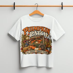 Op de afbeelding: Een wit t-shirt met een grafische print van een vogelverschrikker naast een houten kar vol pompoenen. De tekst luidt PUMPKIN PATCH en FALL HARVEST PUMPKIN PICKING. Het ontwerp bevat herfstbladeren, maïsstengels en een schuur op de achtergrond.