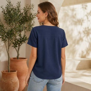 May include: A navy blue short-sleeved blouse with a V-neckline and a straight hemline, paired with light blue denim jeans. The blouse features a simple design with a horizontal seam across the upper back and cuffed sleeves, providing a clean look.
