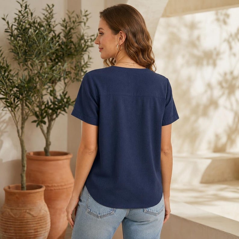 May include: A navy blue short-sleeved blouse with a V-neckline and a straight hemline, paired with light blue denim jeans. The blouse features a simple design with a horizontal seam across the upper back and cuffed sleeves, providing a clean look.