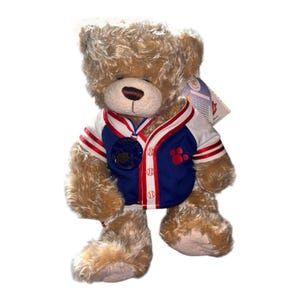Può includere: Un orsacchiotto di peluche con pelliccia marrone chiaro indossa una maglia da baseball blu e bianca con finiture rosse. La maglia presenta una grafica con impronta rossa, bottoni a tema baseball e un medaglione circolare su un nastro tricolore.