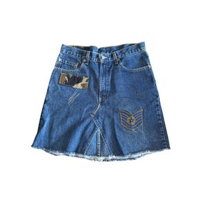 May include: A blue denim skirt with a raw hem. The skirt features a camouflage patch on the front and a military-style patch with a star. The skirt has a classic five-pocket design.