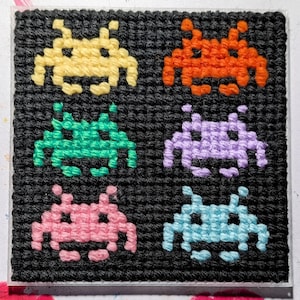 May include: A square, black, crocheted coaster featuring six colourful pixelated alien figures in yellow, orange, green, purple, pink, and blue. The figures are arranged in two rows of three.