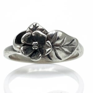 May include: A silver ring featuring a sculpted floral design with a single five-petaled flower and an adjacent leaf. The metal has a polished finish with darkened recessed areas to highlight the intricate details of the petals and the leaf veins.