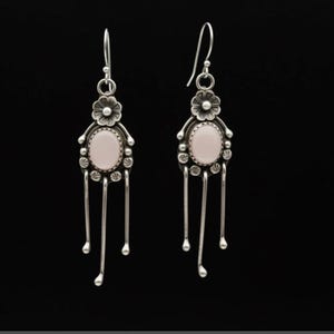 May include: A pair of silver dangle earrings featuring a central oval-shaped pale pink stone. Each earring has a floral motif at the top, a beaded border around the stone, and three thin silver wires hanging down with small rounded tips at the ends.