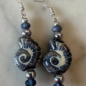 May include: A pair of dangle earrings featuring ceramic ammonite fossil-shaped beads in shades of blue, cream, and brown. Each earring is accented with metallic silver-toned spheres and smaller matte blue beads, suspended from silver-toned ear wires.