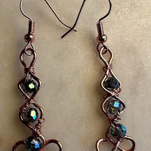 May include: A pair of dangle earrings featuring copper-colored wire wrapped in a decorative, looping pattern. Each earring incorporates three dark, faceted, iridescent beads secured within the wire structure, finished with a standard hook wire.