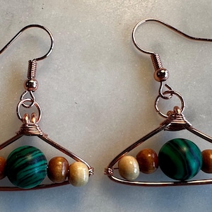 May include: A pair of dangle earrings featuring a triangular copper wire frame. Each earring contains a central green and black marbled bead flanked by two smaller light brown wooden beads and two tan wooden beads on each side.