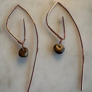 May include: A pair of copper wire earrings featuring a curved, elongated teardrop shape. Each earring is accented with a single, polished, brown and gold tiger's eye gemstone bead suspended from a small coil of copper wire at the base of the frame.