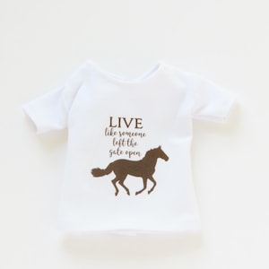 May include: White t-shirt with a brown horse silhouette and the text "LIVE like someone left the gate open". The shirt has short sleeves and a crew neck. The design is centered on the front.