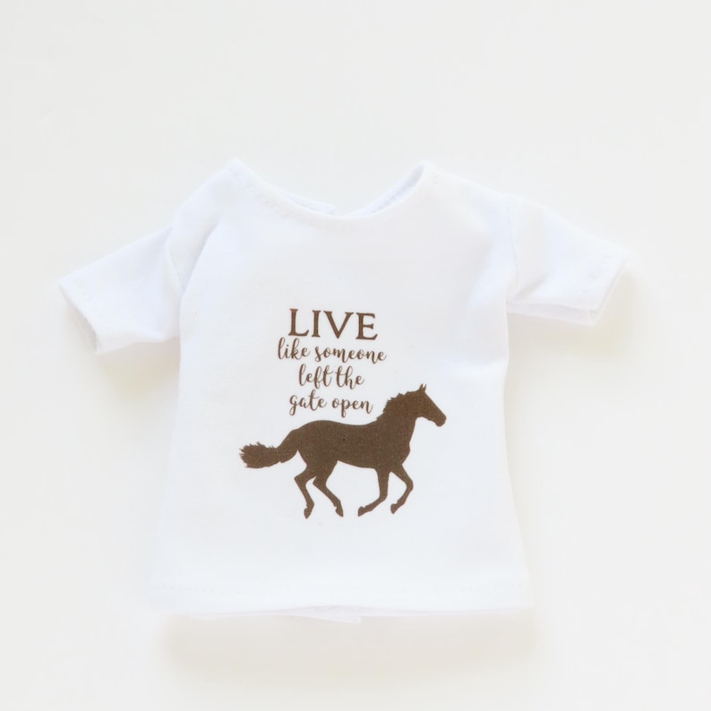 May include: White t-shirt with a brown horse silhouette and the text "LIVE like someone left the gate open". The shirt has short sleeves and a crew neck. The design is centered on the front.