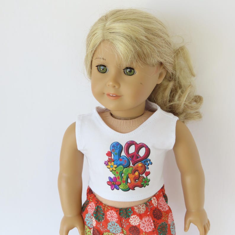 May include: A doll wearing a white crop top with a colorful "Love" graphic and a peace sign. The doll also wears red patterned shorts. The doll has blonde hair and green eyes.