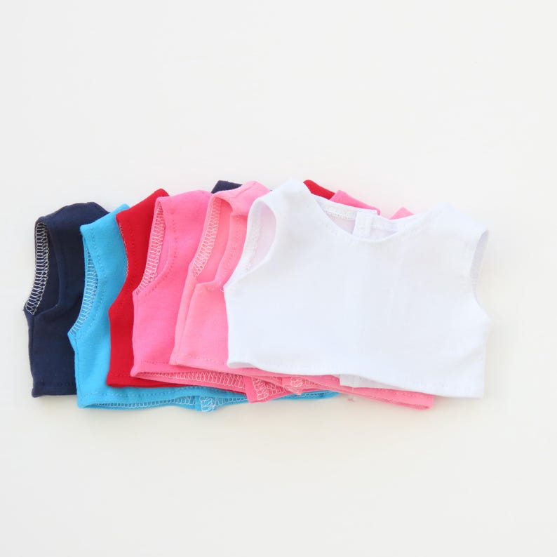 May include: A stack of colorful doll vests in various colors, including navy blue, light blue, red, pink, and white. The vests are sleeveless with a rounded neckline and a small opening in the back.