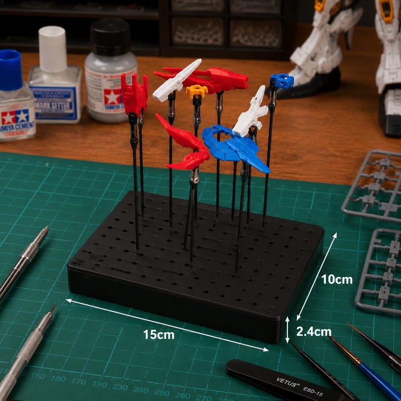 May include: A black plastic model painting stand, 15cm long, 10cm wide, and 2.4cm high, holds various model parts in red, white, and blue. Paint bottles and tools are visible on a green cutting mat, with the text "TAMIYA CEMENT" and "MARK SETTER".