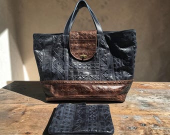 May include: A large, navy blue leather tote bag with a brown leather flap and base, featuring embossed geometric patterns. A matching navy blue clutch is placed in front of the tote bag. The bag has a top handle and a brass clasp.