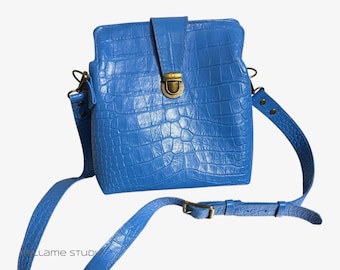 May include: A bright blue crossbody bag with a textured crocodile-embossed pattern. The bag features a rectangular shape, a fold-over flap with a central antique brass-toned metal push-lock closure, and an adjustable matching blue shoulder strap.