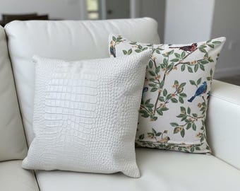 May include: Two decorative throw pillows on a white sofa. One pillow is white with a crocodile skin texture. The other pillow has a cream background with a pattern of birds, leaves, and flowers in various colors.