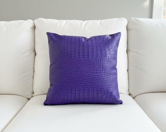 May include: A square, purple decorative pillow with a faux alligator skin texture. The pillow is placed on a white sofa, with a white pillow behind it. The pillow is a vibrant shade of purple.