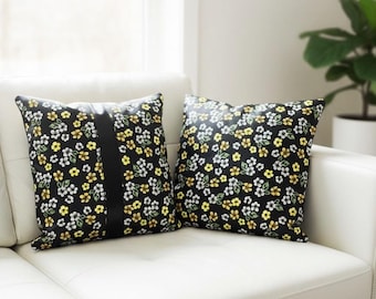 May include: Two square throw pillows with a black background and a floral pattern of yellow, white, and green flowers. One pillow has a vertical black stripe down the center. The pillows are on a white couch.