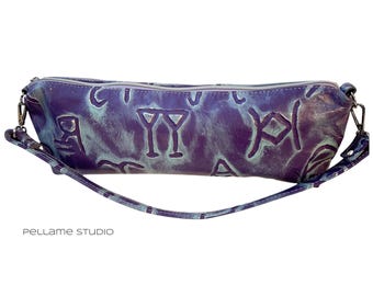 May include: A long, cylindrical, purple and teal leather bag with a zipper closure and a detachable strap. The bag features embossed symbols across its surface. The words "pellame studio" are printed in white at the bottom.