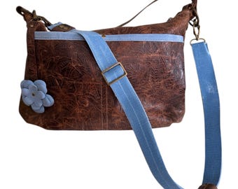May include: A brown shoulder bag with a textured floral pattern, featuring a light blue fabric trim and a matching light blue adjustable crossbody strap. A light blue flower-shaped embellishment is attached to the front, and the text PELLAME STUDIO is printed at the bottom.
