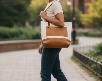 May include: A tote bag featuring a cream-colored canvas upper section and a tan leather lower section. The bag includes a tan leather exterior pocket with a horizontal zipper closure, brown leather shoulder straps, and metal hardware attachments.