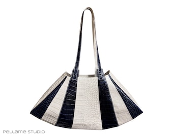 May include: A structured tote bag featuring a fan-shaped silhouette with alternating vertical panels of cream and dark navy blue leather. The material has a textured crocodile-embossed finish. It includes two long shoulder straps and the text PELLAME STUDIO.