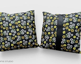 May include: Two square throw pillows with a black background and a floral pattern in yellow, white, and green. One pillow has a vertical black stripe. The pillows are on a white background.