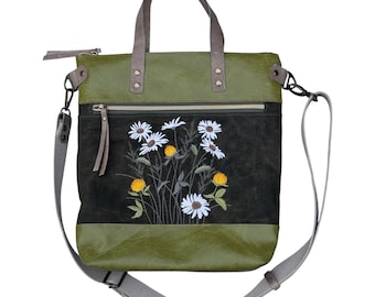 May include: A green and black tote bag featuring an embroidered floral design of white daisies and yellow clover blossoms. The bag includes two top handles, a detachable gray shoulder strap, a zippered top closure, and a front zippered pocket.